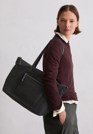 Frau trägt einen bordeauxfarbenen Pullover und graue Hose und hält eine große schwarze Tote-Bag mit einer Reißverschlusstasche außen über der Schulter.