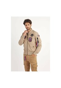 Veste bomber beige avec des zips, des badges et des franges colorées. Matériau léger, poignets et taille côtelés pour une silhouette ajustée.