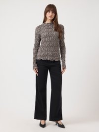 Femme debout portant un haut long-sleeve texturé gris, un pantalon large noir et des escarpins noirs à bout pointu sur un fond blanc.