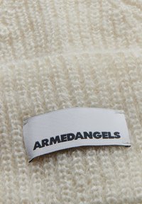 Dik romig gebreid stof met een zachte textuur en zichtbare steken. Bevat een wit label met "ARMEDANGELS" in zwarte letters.