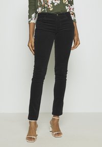 Kvinna som bär svarta slim-fit byxor, en blommig grön topp och vita öppna klacksandaler med blockklack, stående mot en enkel ljus bakgrund.