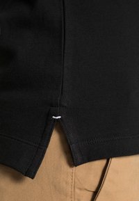 Dobladillo de tela texturada negra de una camisa con una pequeña abertura lateral y detalle de costura blanca sobre pantalones beiges.