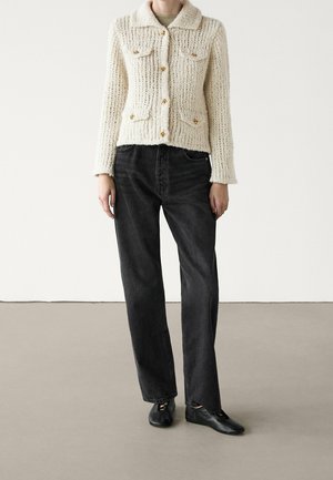 Personne portant une veste en maille crème boutonnée avec des boutons dorés, un jean noir taille haute droit et des chaussures plates noires, debout sur un sol gris.