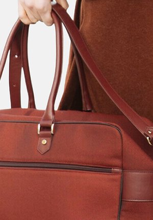 Personne tenant un sac de voyage avec poignée en cuir marron, doté d'une poche avant zippée, sur un fond en tissu marron.