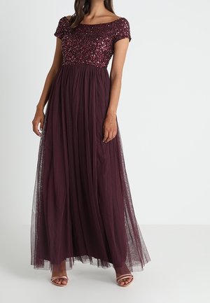 Kobieta ubrana w długą, sięgającą podłogi suknię w ciemnym fiolecie z cekinowym gorsetem z krótkim rękawem i spódnicą z tiulu, do której ma dopasowane sandały na otwartym obcasie.