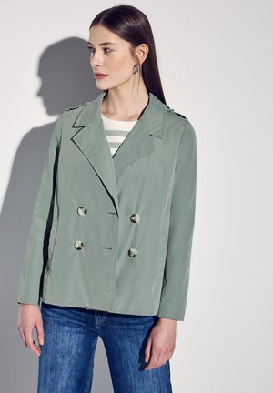 Veste double boutonnage vert clair avec de gros boutons, épaulettes, et manches longues, associée à un pull rayé et un jean bleu.