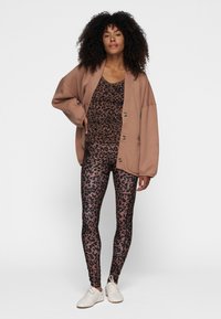Cárdigan marrón de gran tamaño sobre una camiseta sin mangas negra con estampado de leopardo y leggings. Los leggings tienen una textura brillante y se combinan con zapatillas blancas.
