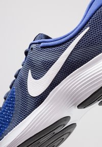 Zapato deportivo con una parte superior de malla azul marino, acentos azules, logo en forma de swoosh blanco y una media suela texturizada blanca. Suela de goma negra con patrones de agarre.