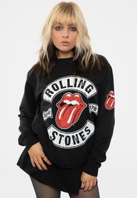 Black long-sleeve sweatshirt featuring Rolling Stones logo in red and white, with vintage textured graphics and a small logo patch on the right sleeve.