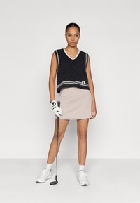 Námořnická pletená vesta s bílým lemem přes béžovou skládací sukni. Modelka drží golfovou hůl a má na sobě bílé tenisky a bílou golfovou rukavici.