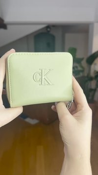 Zaļa mākslīgās ādas maks ar reljefa "cK" logotipu, rāvējslēdzēja aizdari un taisnstūra formu, ar redzamu šuvumu un gludu tekstūru.