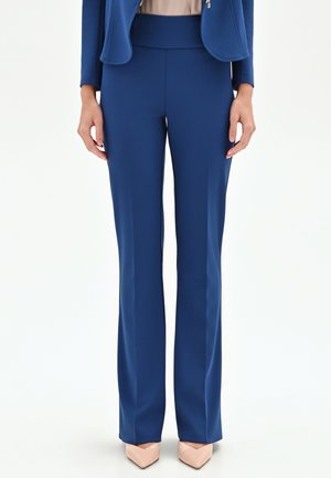 Femme portant un pantalon bleu taille haute à jambes droites assorti à un blazer bleu et des escarpins nude à bouts pointus, debout devant un fond blanc.