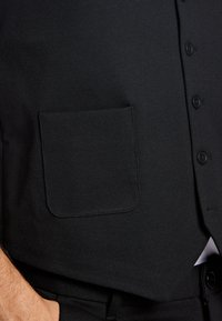 Schwarze Weste aus strukturiertem Stoff, mit einer Fronttasche und Knopfverschlüssen. Das Design ist tailliert und angepasst.