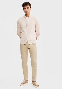 Jasno beżowa kurtka na zamek, pod nią biała koszula, w zestawie z beżowymi spodniami i brązowymi trampkami. Gładka tekstura, swobodny krój, bez wyrazistych wzorów.