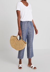 Bílý tričko s výstřihem do V, světle modré lněné zkrácené kalhoty, tkaná slaměná kabelka a bílé sandály se dvěma popruhy, stojící na světlém pozadí.