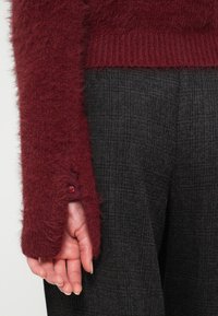 Bordo puhasti pulover z rebrastimi manšeti, ki ima gumbasto podrobnost na rokavu. V kombinaciji z teksturiranimi sivimi hlačami.