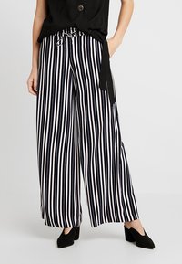 Svarta och vita randiga vida byxor med dragsko i midjan och slät tyg, tillsammans med svarta spetsiga klackskor.