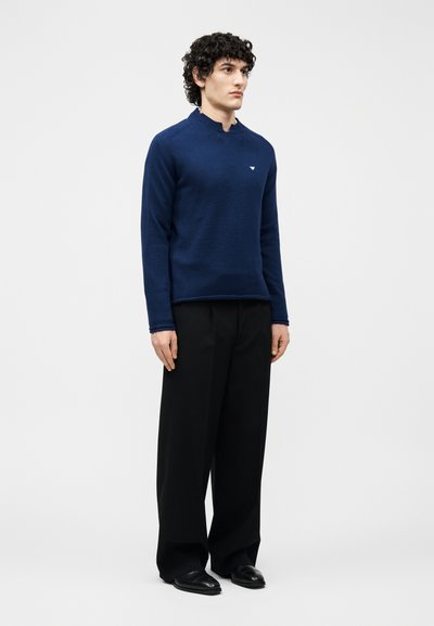 Άνδρας που φορά φαρδιά μαύρη παντελόνια, μαύρα παπούτσια και εφαρμοστό μπλε ναυτικό πουλόβερ με μικρό λευκό λογότυπο στο στήθος.