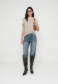 Pull beige en maille avec encolure ronde, associé à un jean bleu clair à coupe ample. Bottes en cuir foncé montant jusqu'aux genoux. Sac à épaule marron. Lunettes transparentes.