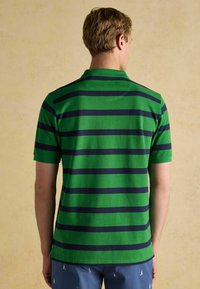 Zaļš un jūras zils svītrains polo krekls ar īsām piedurknēm, ribotu apkakli un sānu ventiļu detaļu. Mīksta, gluda auduma ar ikdienišķu piegriezumu.