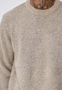 Personne portant un pull en maille beige à texture, avec un cou tatoué visible présentant des motifs de roses et de lettres.