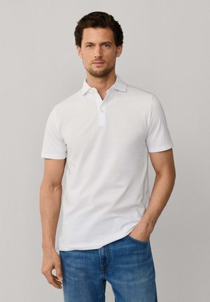 Mann mit kurzen dunklen Haaren, der ein weißes kurzärmeliges Poloshirt und blaue Jeans trägt und vor einem schlichten hellen Hintergrund steht.