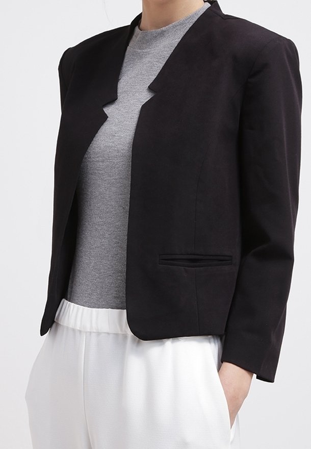 Femme portant un blazer noir cintré sur un haut gris et un pantalon blanc, avec une main dans la poche, sur un fond uni.