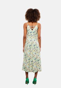 Blumiges Midikleid mit Spaghettiträgern, hellgrünem Stoff, blauen und gelben Blumenmustern sowie einem gekreuzten Rückendetail. Kombiniert mit grünen Absatzschuhen.
