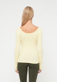 Pitkähihainen ribbipaita pehmeässä keltaisessa, jossa on leveä avonainen selkä, tiukka muotoilu ja pitkät hihansuut, yhdistettynä oliivinvihreisiin housuihin.