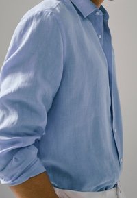 Vaaleansininen pellavapaita, jossa on napillinen kaulus. Ominaisuuksiin kuuluu käärityt hihat ja rento istuvuus. Sileä tekstuuri ja hienovarainen kiilto.