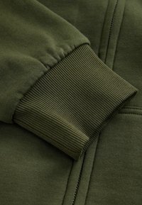 Olivengrøn sweatshirt med ribbede manchetter, blød tekstur og lynlås foran. Nærbillede af manchetdetaile med et glat, let strækbart stof.