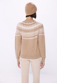Person mit beigefarbenem, gemustertem Strickpullover und passender Mütze, kombiniert mit hellbeigen Hosen, steht mit dem Rücken gegen einen weißen Hintergrund.