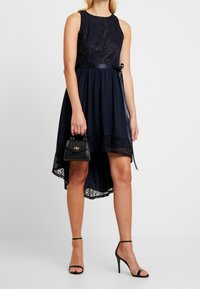 Robe bleu marine avec un corsage en dentelle, jupe évasée et ourlet asymétrique. Présente une taille en satin et est accompagnée d'un petit sac à main noir.