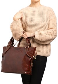 Borsa a tracolla in pelle marrone con manici doppi, con accenti in ottone e cuciture. Maglione beige intrecciato indossato da una persona che tiene la borsa.