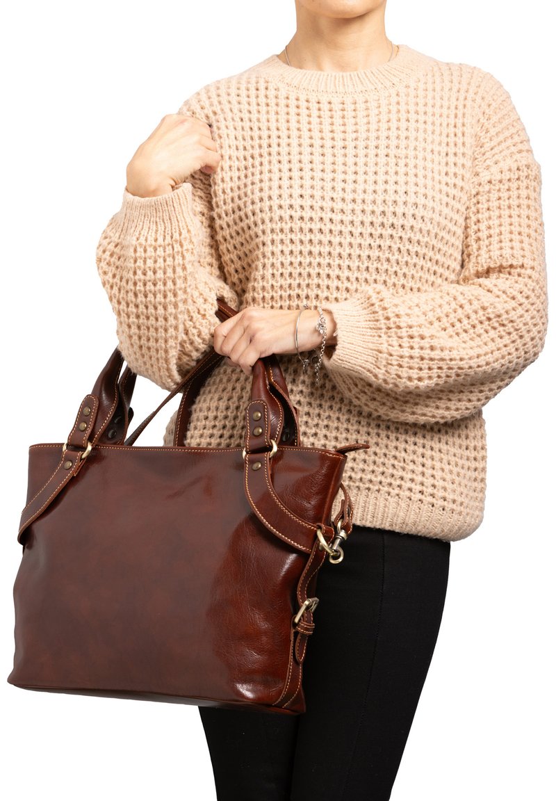 Borsa a tracolla in pelle marrone con manici doppi, con accenti in ottone e cuciture. Maglione beige intrecciato indossato da una persona che tiene la borsa.