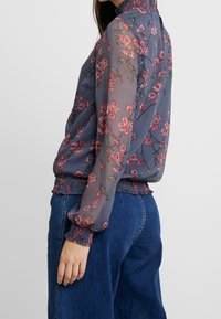 Průhledná květinová blůza v námořnické modři s růžovými květy; vysoký límec, elastické manžety a lem. V kombinaci se volnými modrými džíny.