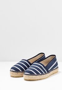 Marinblå canvas-espadriller med vita ränder, rund tå och sulan omsluten av jute. Har en platt, flexibel design med en bekväm öppning.
