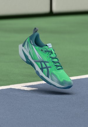 Scarpa da tennis Asics verde brillante e teal che fluttua sopra linee del campo blu e verde.