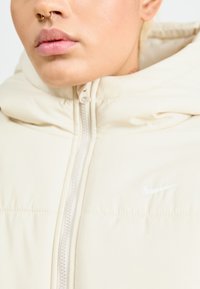 Casaco acolchoado de cor bege clara com gola alta, fecho de correr e um logótipo branco da Nike na frente. Textura suave, tecido liso.