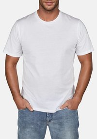 Weißes Baumwoll-T-Shirt mit Rundhalsausschnitt und kurzen Ärmeln, präsentiert an einem Modell, das Jeans trägt und die Hände in den Taschen hat.