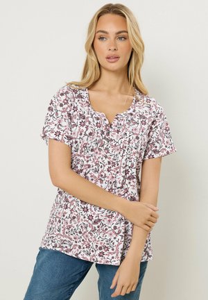 Frau mit langen blonden Haaren, die eine kurzärmelige weiße Bluse mit lila Blumenmuster und blaue Jeans trägt, steht vor einem einfarbigen Hintergrund.