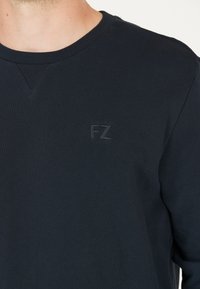 Mörk marinblå sweatshirt med rund hals och strukturerad tyg. Har en liten, tonad "FZ"-logga på bröstet. Mjuka, långa ärmar.