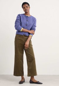Lila stickad cardigan med rund hals, tre knappar och långa ärmar; parat med vida olivgröna kordbyxor och mörka platta skor.