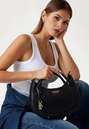 Borsa a mano in pelle nera con dettagli intrecciati, logo dorato e charm. Presenta doppi manici e una silhouette curvata. Abbinata a una maglietta bianca e jeans.