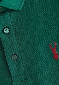 Polo verde con tessuto testurizzato, caratterizzato da tre bottoni e un logo ricamato di un cervo rosso sul lato sinistro.