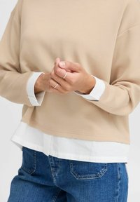 Beige sweatshirt met een ontwerp met een verlaagde schouder, gelaagd over een wit overhemd met knopen. Gedragen met blauwe spijkerbroek, met zilveren ringen aan de vingers.