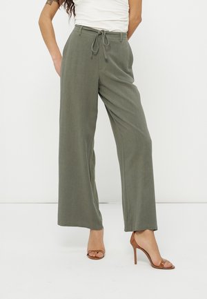 Femme portant un pantalon large taille haute vert olive avec cordon et des sandales à talons marron, debout sur fond blanc.