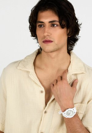 Orologio in silicone bianco con cassa rotonda e accenti argentati, abbinato a una camicia beige chiaro con texture e colletto a bottoni.