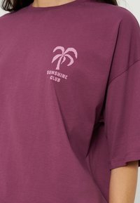 Femme portant un t-shirt violet avec un petit palmier rose et le texte "Sunshine Club" sur la partie supérieure gauche de la poitrine.