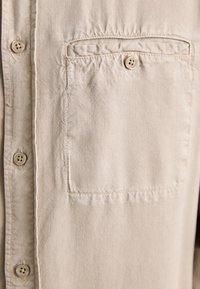 Beige nappipaita, jossa on rintatasku ja pehmeä, teksturoitu kangas näkyvine ompeleineen taskun ja nappien ympärillä.